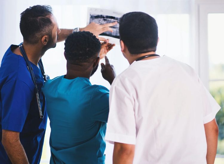 Three medical professionals in scrubs and lab coats closely examining an X-ray film. One person points to a specific area on the image, indicating active discussion. They stand in a bright clinical room with large windows, suggesting a hospital or diagnostic setting.