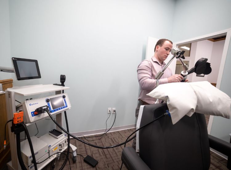A clinician adjusts a Transcranial Magnetic Stimulation (TMS) device in a treatment room. The setup includes a mechanical arm holding a magnetic coil positioned near a padded chair, a control unit with a digital display, and a computer monitor. The environment reflects a professional and modern approach to administering TMS protocols for neurological and psychiatric care.
