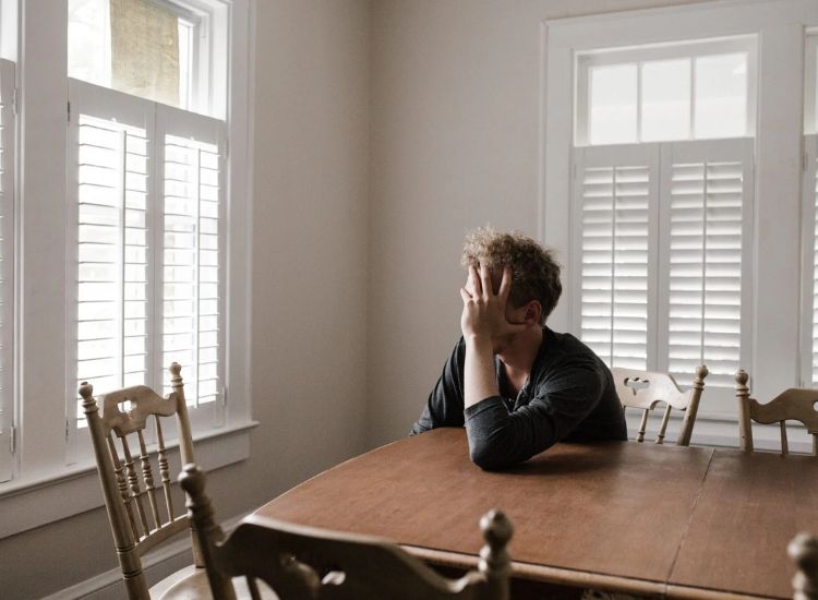 A person sits alone at a wooden dining table in a naturally lit room, resting their head on one hand in a contemplative pose. The space features white walls, plantation shutters, and four wooden chairs, evoking a quiet moment of introspection — visually representing the emotional nuance often misunderstood in depression narratives.