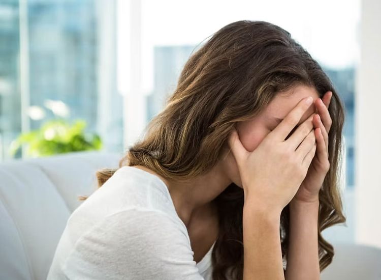 Person sitting indoors with long brown hair, wearing a white top, covering their face with both hands in a moment of distress.