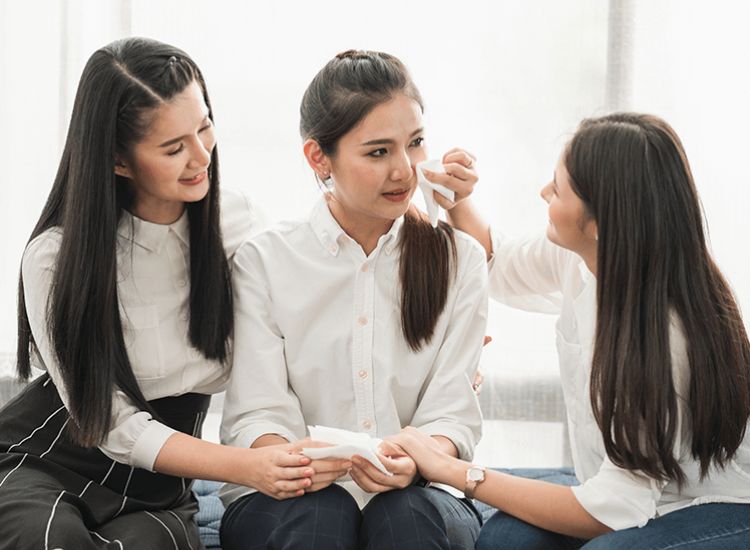 Three people indoors offering emotional support: two individuals comfort a crying person in the center by holding hands and gently wiping tears with tissues.