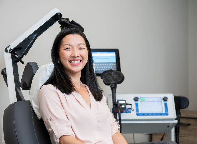 A person seated in a clinical setting undergoes Transcranial Magnetic Stimulation (TMS), with a circular coil positioned near their head and a digital control unit nearby, illustrating a non-invasive brain stimulation procedure used in mental health treatment.