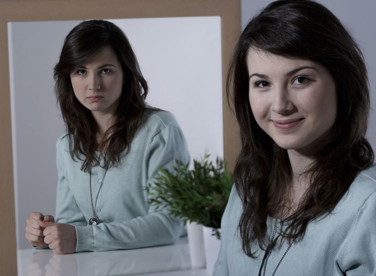 Woman in a light blue cardigan smiling at the camera, while her mirror reflection shows a serious expression; a small green potted plant sits on the table in front.
