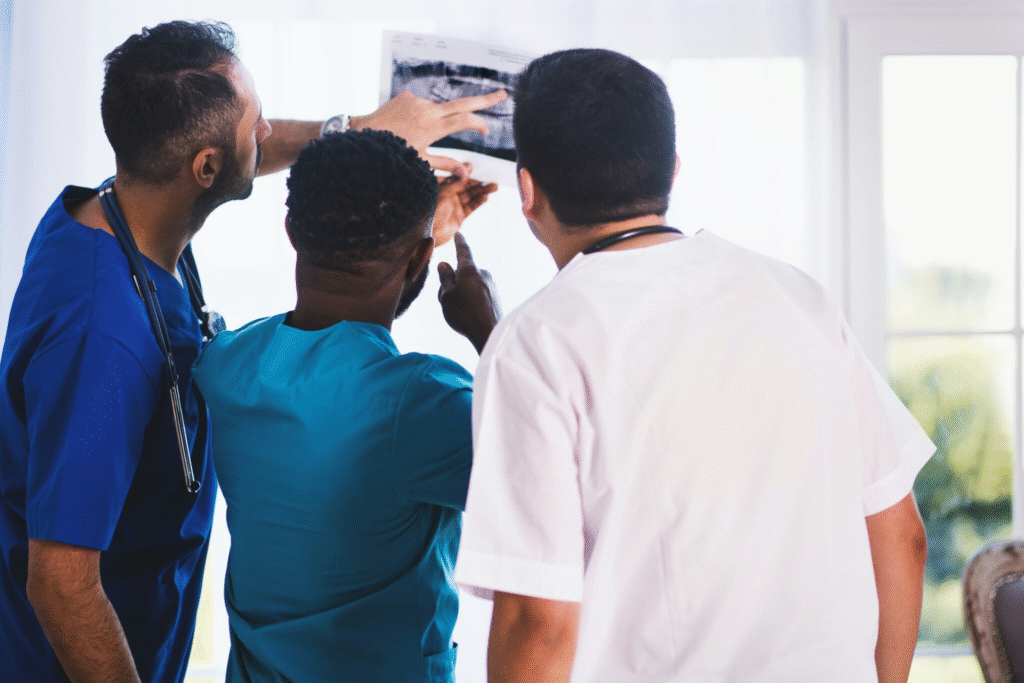 Three medical professionals in scrubs and lab coats closely examining an X-ray film. One person points to a specific area on the image, indicating active discussion. They stand in a bright clinical room with large windows, suggesting a hospital or diagnostic setting.