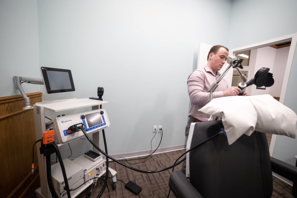 A clinician adjusts a Transcranial Magnetic Stimulation (TMS) device in a treatment room. The setup includes a mechanical arm holding a magnetic coil positioned near a padded chair, a control unit with a digital display, and a computer monitor. The environment reflects a professional and modern approach to administering TMS protocols for neurological and psychiatric care.