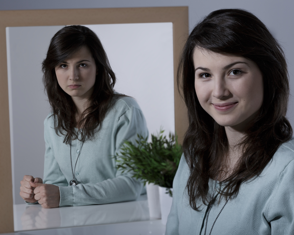 Woman in a light blue cardigan smiling at the camera, while her mirror reflection shows a serious expression; a small green potted plant sits on the table in front.